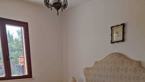 A bedroom with a wooden floor, a bed with a floral headboard, a bedside table with a lamp, and a window with a view of greenery. - Villa sul Mare (Sorso)