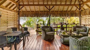 A covered outdoor seating area with wicker furniture, a wooden deck, and a view of palm trees. - Fare Oaoa · on the Haapiti surf pass, sleeps 4 (Moorea)