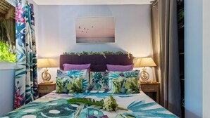 A bedroom with a bed, floral curtains, a headboard with greenery, a painting of birds, and two bedside lamps. - Fare Oaoa · separate living room, beachfront, sleeps 4 (Moorea)