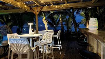 Eine Terrasse im Freien mit weißen Korbstühlen und -tischen, Lichterketten und Blick auf Palmen und das Meer.