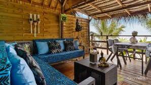 A wooden cabin with a thatched roof, a blue patterned sofa, a dining table, and a view of palm trees. - Fare Oaoa · separate living room, beachfront, sleeps 4 (Moorea)