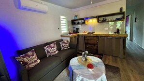 A compact living space with a sofa, a small table with a vase, a ceiling fan, a kitchenette, and a window with blinds. - Fare Oaoa · own screened terrace, sleeps 4 (Moorea)