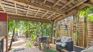 Een overdekt terras met houten vloer, een tafel, stoelen en een rieten bank.