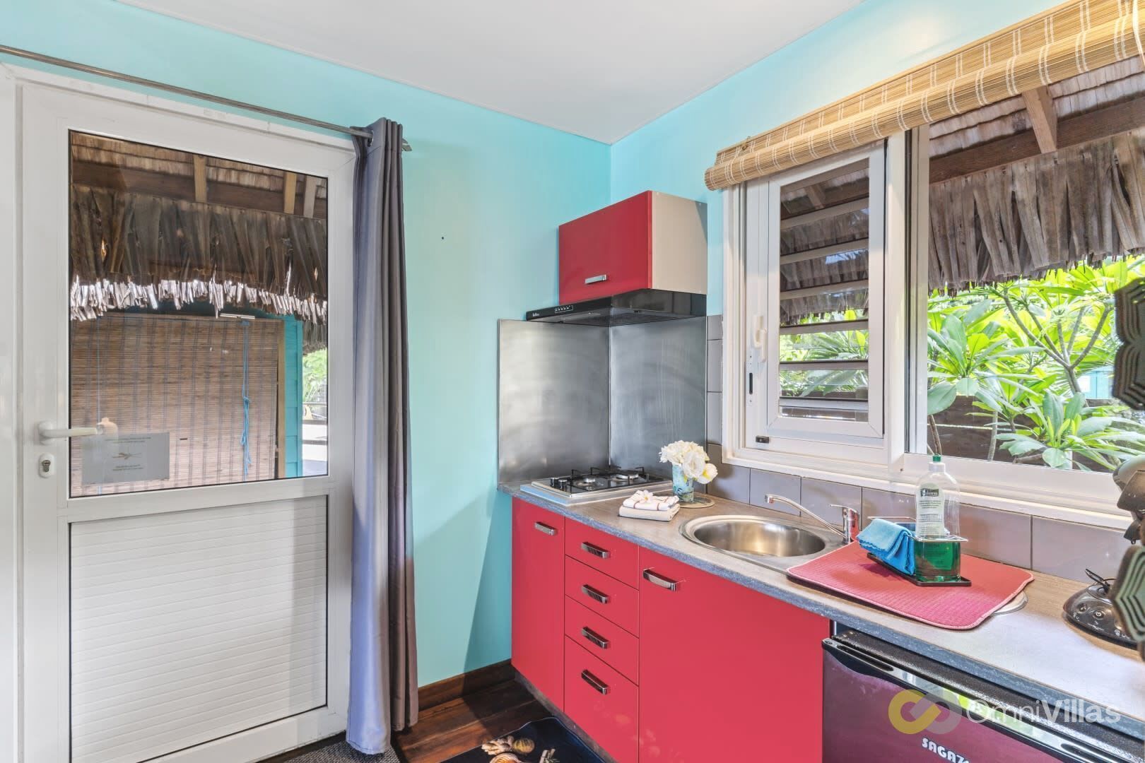 A compact kitchen with a red countertop, stainless steel sink, and a window with a view of greenery.
