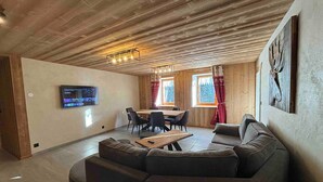Una sala de estar acogedora con techo de madera, un sofá modular grande, una mesa de comedor con sillas y un televisor de pantalla plana montado en la pared.