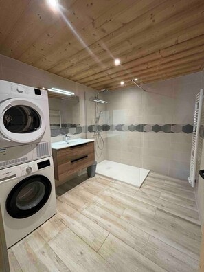 Una lavanderia con lavatrice e asciugatrice affiancate, soffitto in legno e zona doccia con pareti e pavimento piastrellati.