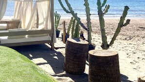 Un espai a la platja amb zona de seients, que inclou un toldo blanc i coixins, tot envoltat de cactus en testos.