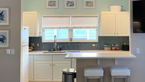 Una cocina moderna con armarios blancos, un lavamanos de acero inoxidable y una ventana con persianas. Hay tres cuadros enmarcados en la pared sobre la ventana.