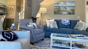Una sala de estar con un sofá azul, un sillón estampado y una mesa de centro de vidrio.