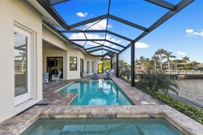 A swimming pool with a glass-enclosed patio area, a covered seating area, and a view of palm trees and a body of water.