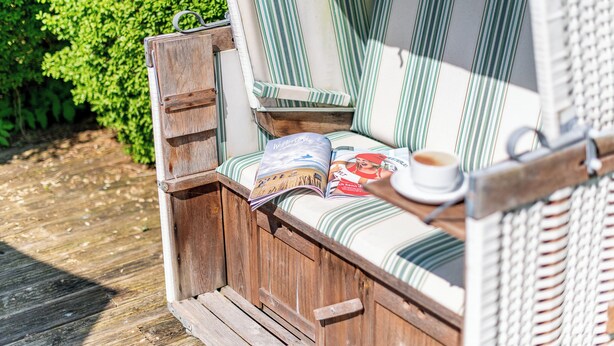 Eine Holzterrasse mit einer Bank und einem Korbsessel, einer Zeitschrift und einer Kaffeetasse.