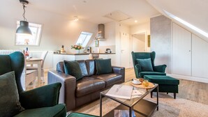 Ein modernes Wohnzimmer mit einer Couch, Sesseln und einem Couchtisch. Im Hintergrund ist eine Küchenzeile mit einem Fenster und einem Oberlicht zu sehen.
