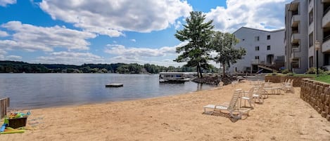 A beach with lounge chairs and a boat on the water, a building with balconies, and a clear sky.