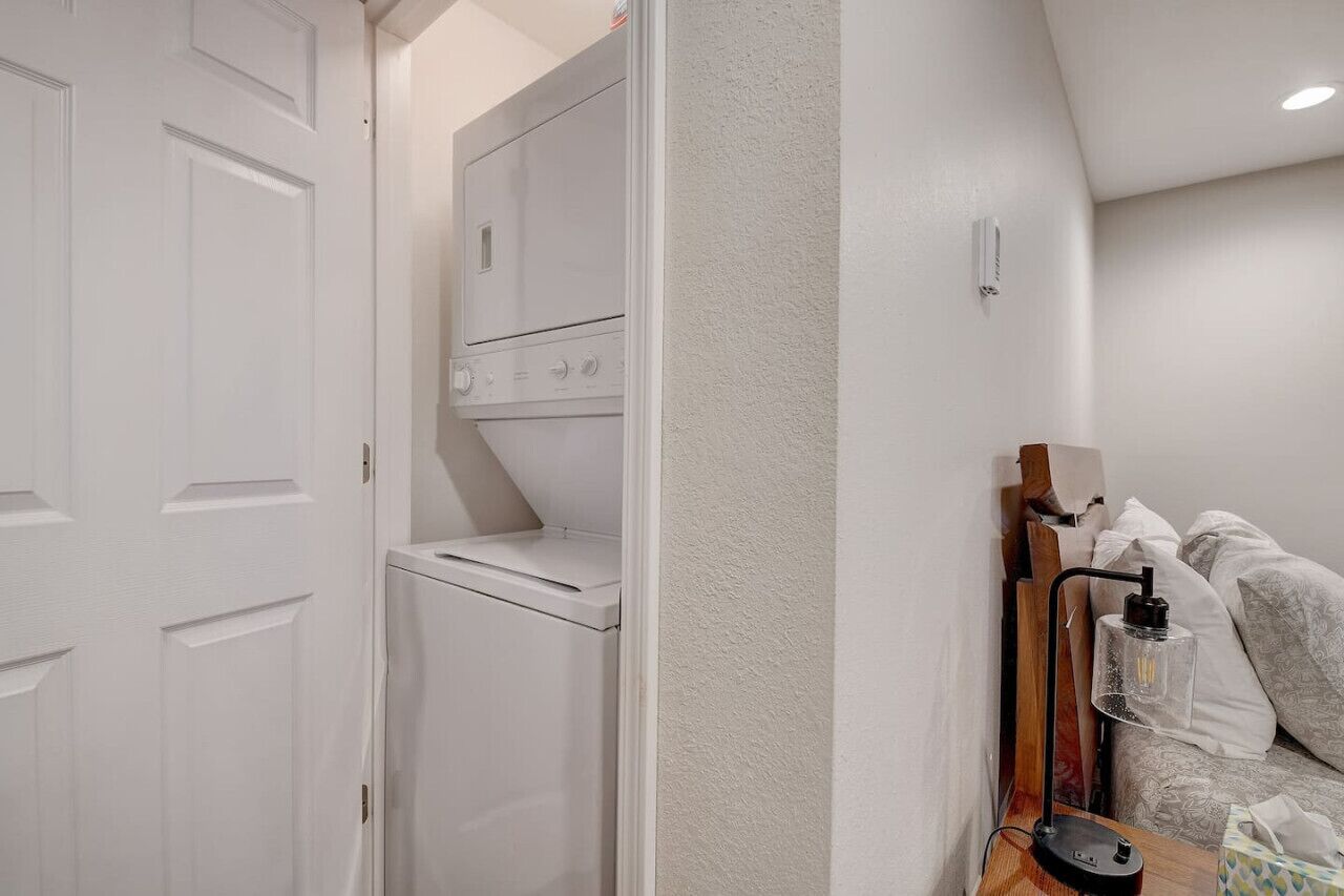 A laundry room with a washing machine and dryer, a wooden headboard with a lamp, and a bedside table with a tissue box.