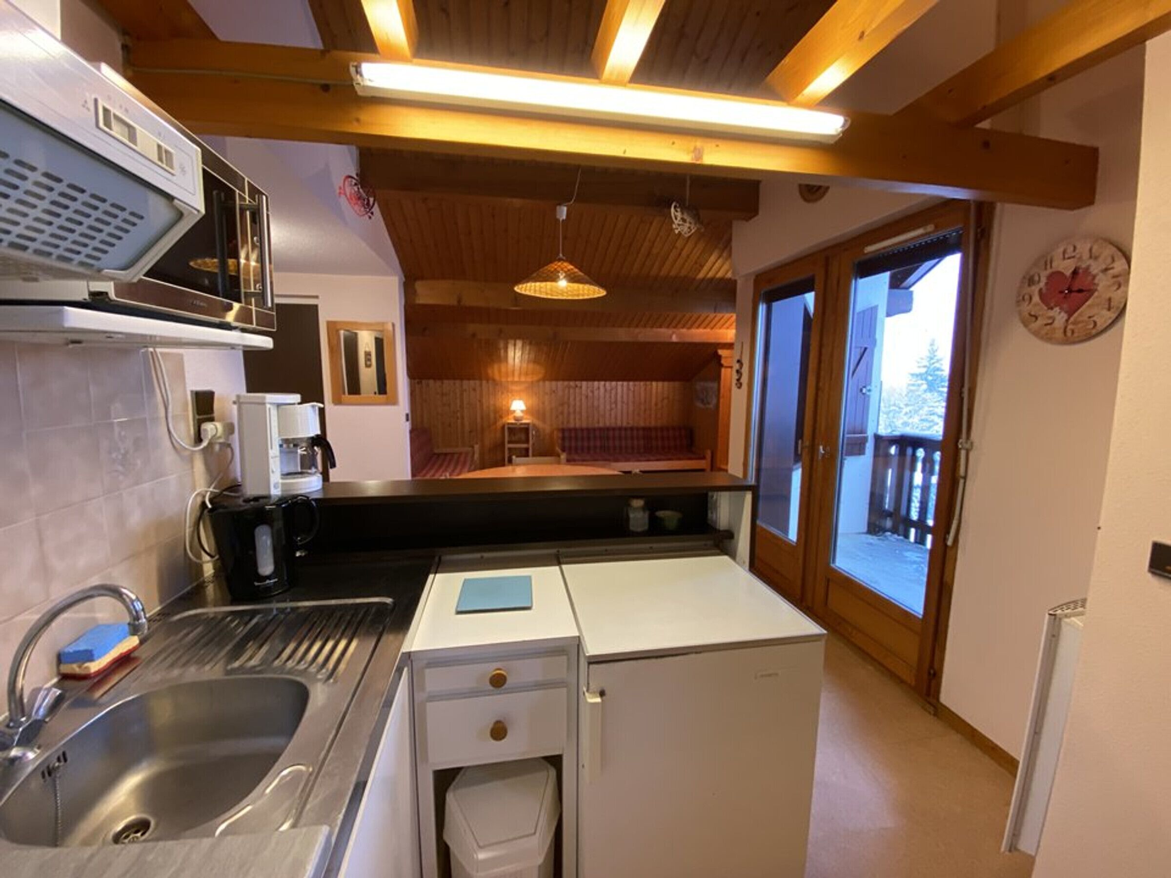 Une cuisine compacte équipée d’un évier, d’une machine à café et d’un petit réfrigérateur. Le plafond est en bois, avec des poutres apparentes, et un luminaire suspendu. Une fenêtre offre une vue sur l’extérieur.