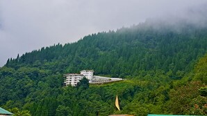 Un edificio de varios pisos con techo inclinado, rodeado de un bosque frondoso y montañas al fondo.