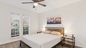 Une chambre à coucher avec un lit, deux tables de chevet, un ventilateur de plafond et une fenêtre avec des stores.