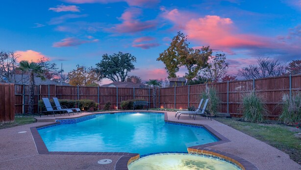 Une piscine rectangulaire avec un jacuzzi, entourée d’une clôture en bois, et un espace patio aménagé avec des chaises longues.