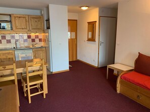 A compact kitchen with wooden cabinets, a microwave, and a refrigerator. There is a dining table with chairs and a small bench with a red cushion. - Cozy apartment in Orcières with parking and free WIFI, 4 people (Orcières Merlette)