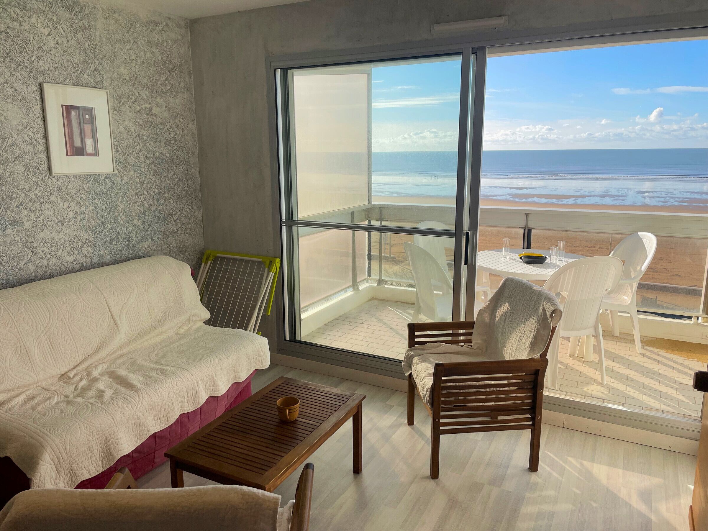 A room with a large window overlooking a beach, a sofa, a wooden table, and two chairs.