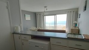 A compact kitchen with a dining area, a view of the beach, and a small table with a lamp.