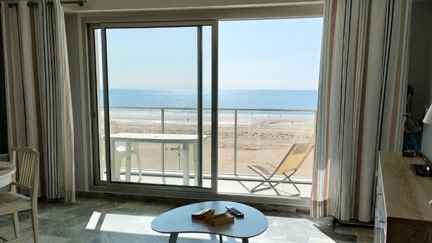 A room with a large sliding glass door leading to a beach view, a small round table with a book and remote control, a beige sofa, and a chair.