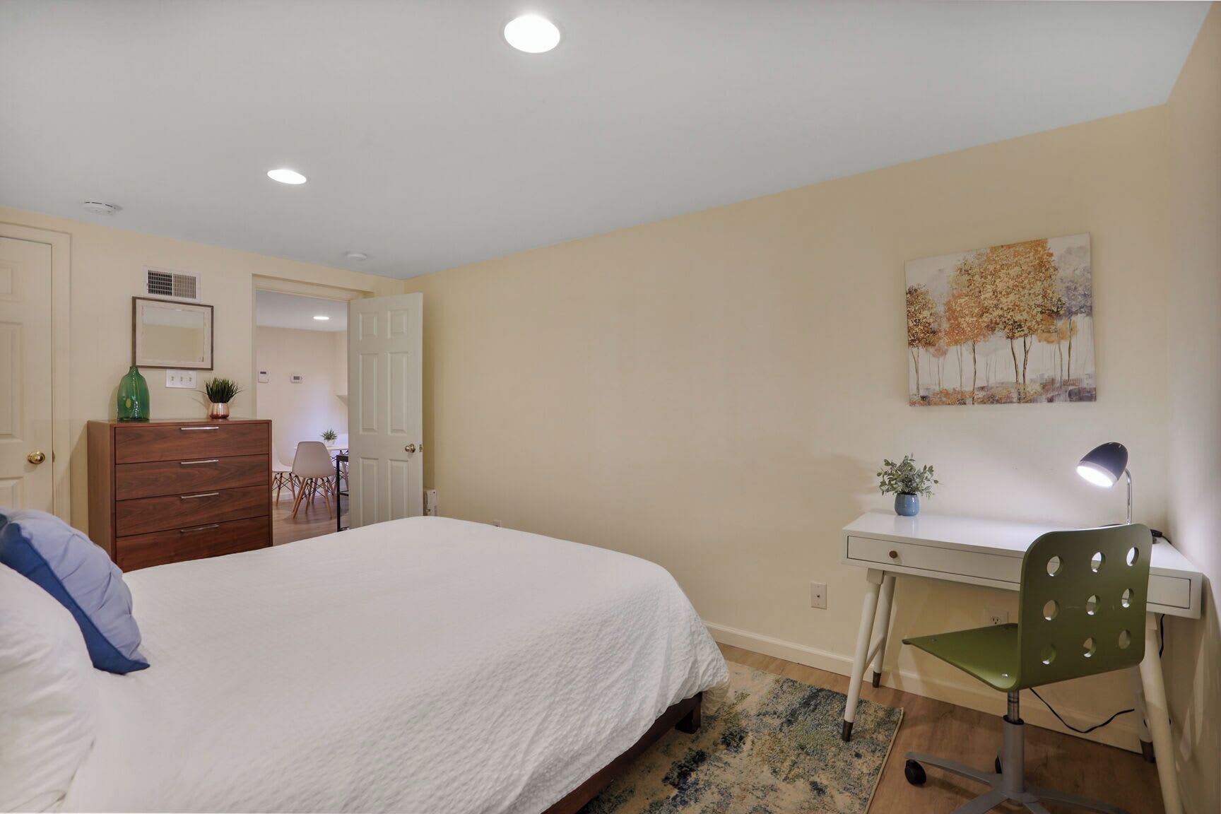 A bedroom with a bed, dresser, desk, chair, and a painting on the wall.