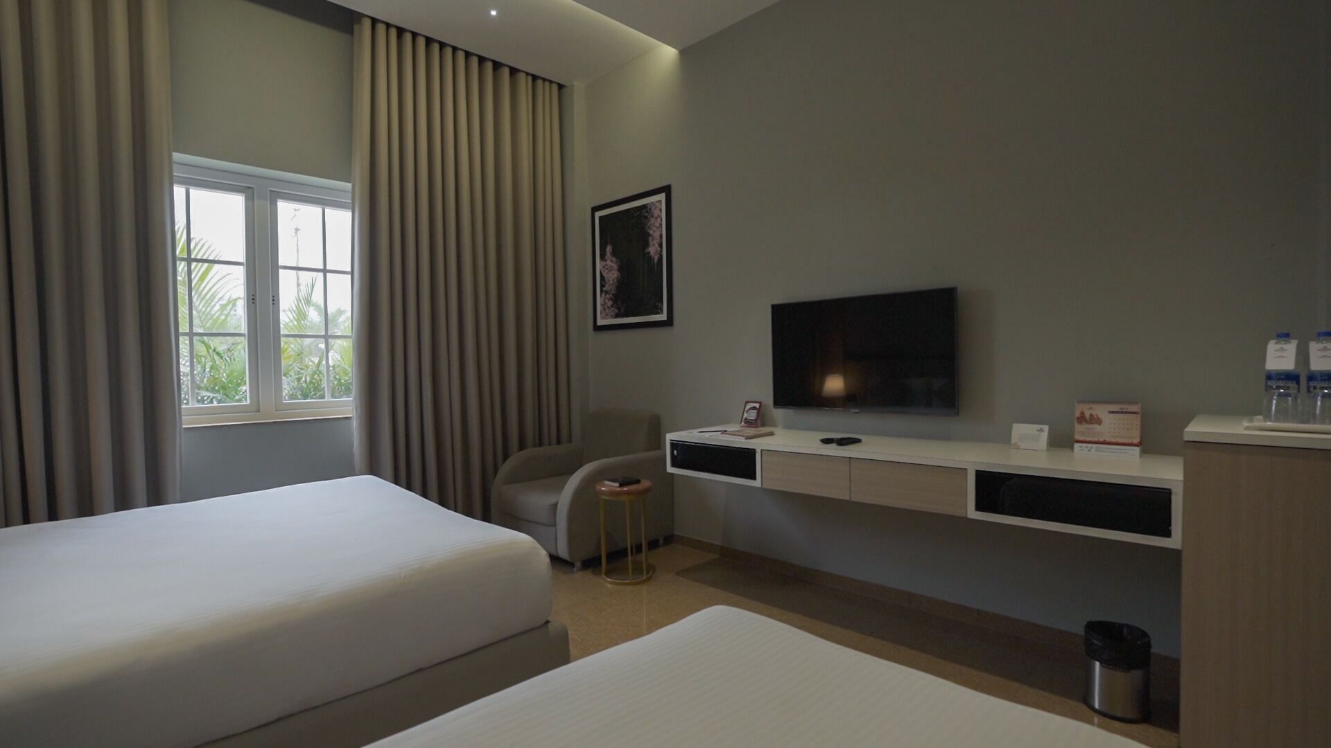 Habitación de hotel con dos camas, un escritorio con televisor, una silla y un ventanal con cortinas.