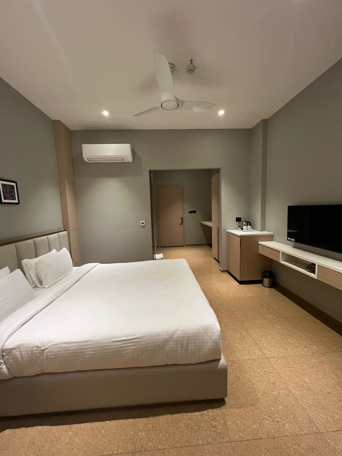 A modern hotel room with a large bed, a TV, a ceiling fan, and a small kitchenette.