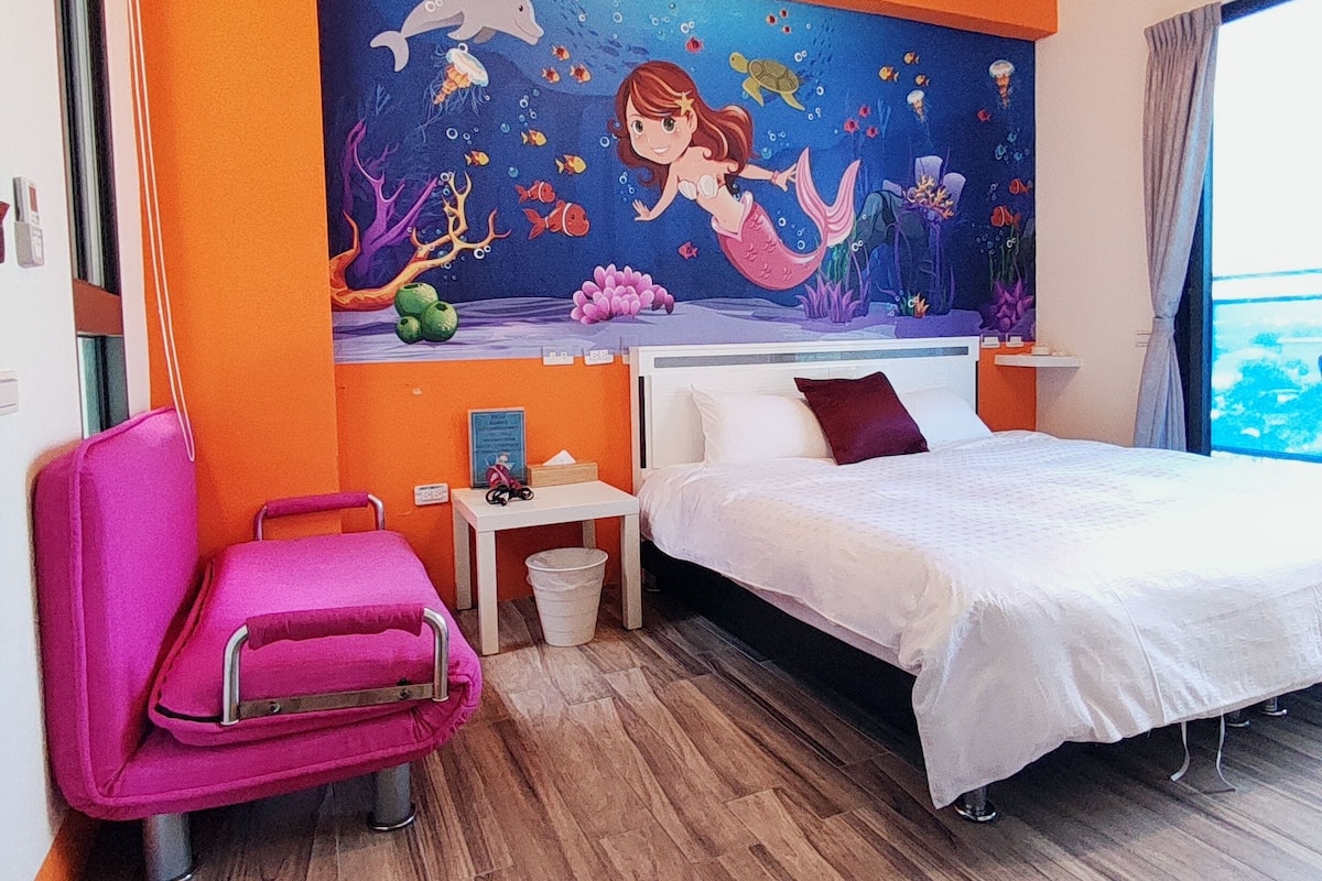 A hotel room with a bed, a chair, a nightstand, and a mural of a mermaid.
