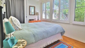 Un dormitorio con cama, mesita de noche, lámpara, ventana con persianas y vista a la vegetación.