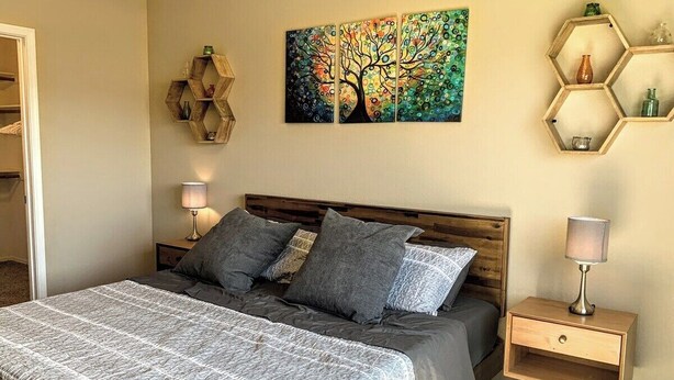 A bedroom with a bed, a nightstand, a lamp, a wall art piece, and a wooden shelf. - Thunder Ridge (Tucson)