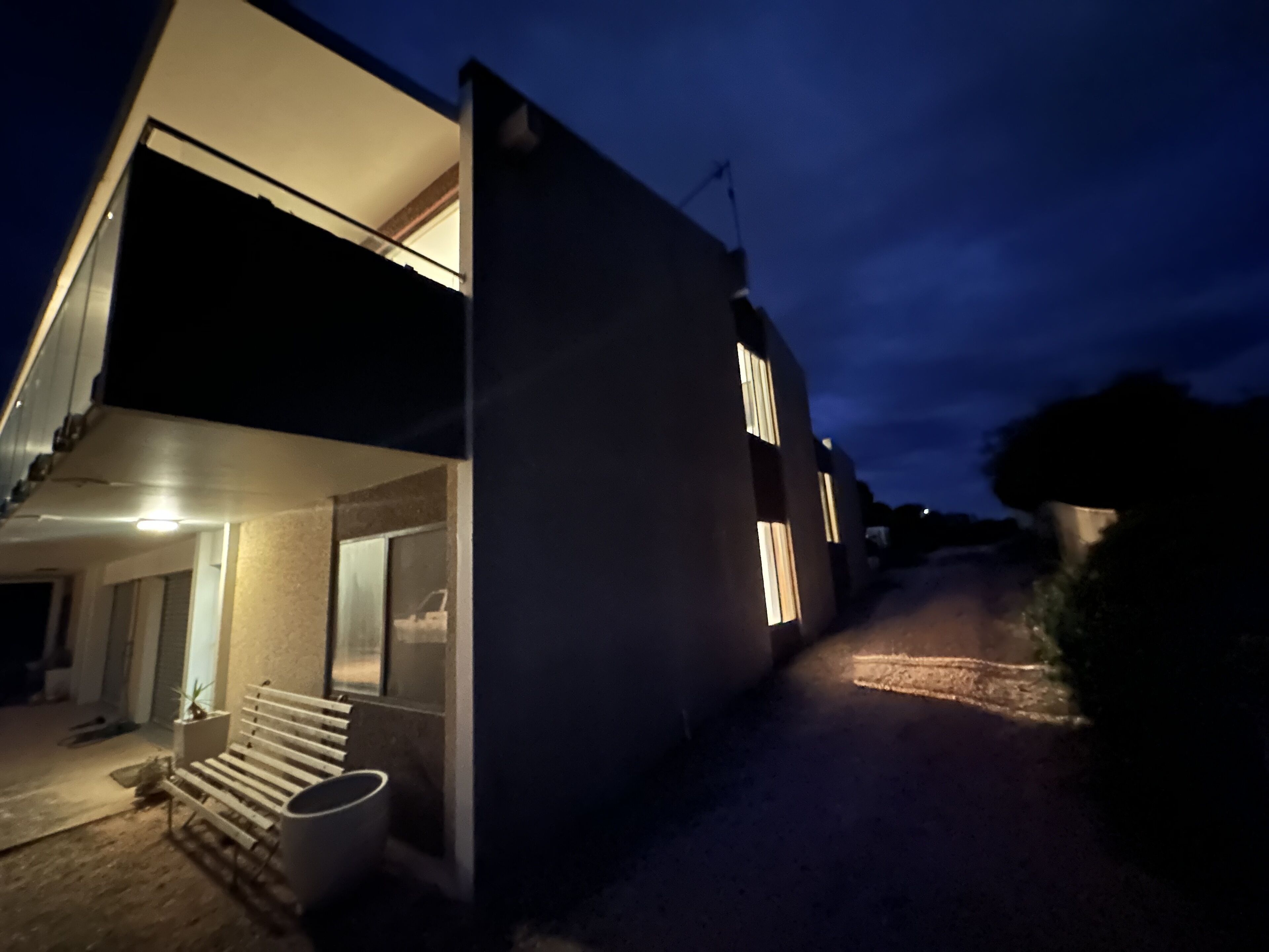 A modern two-story house with a flat roof and large windows. There is a staircase leading to the upper floor, and a small outdoor seating area with a bench and a table.