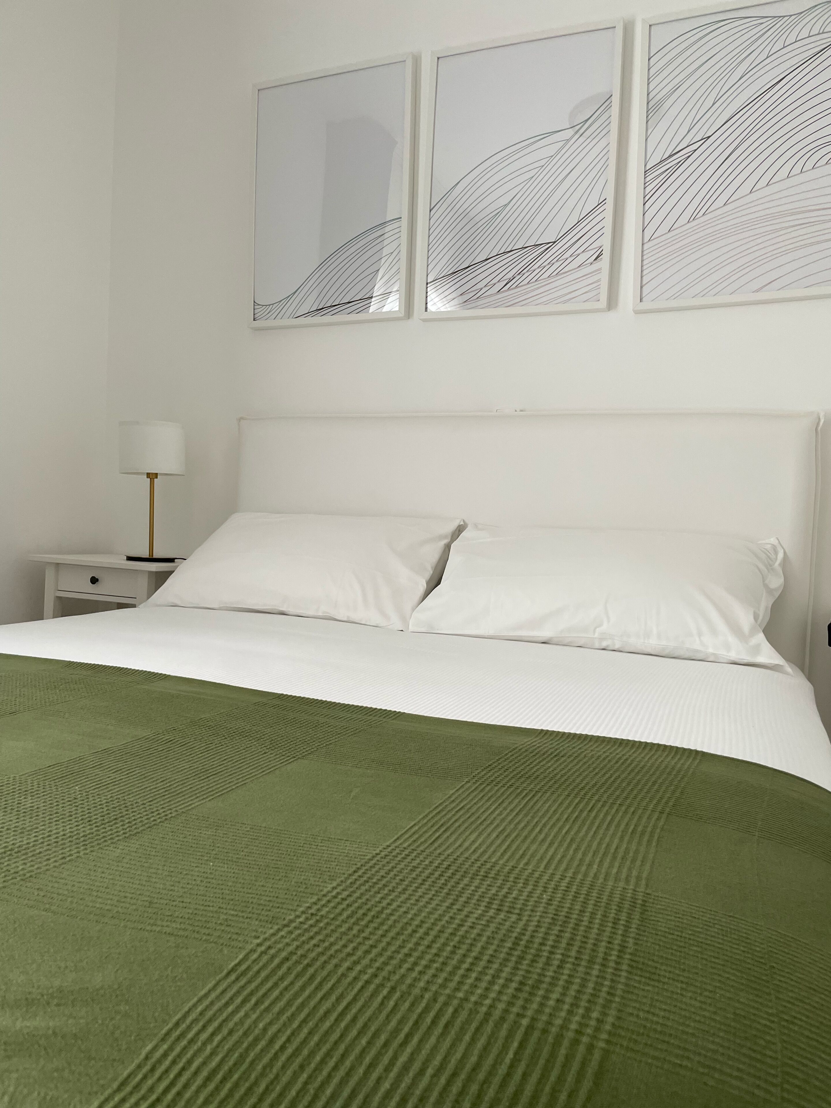 Uma cama bem arrumada com lençóis brancos e um cobertor verde, uma mesa de cabeceira branca com uma luminária, e três obras de arte abstratas na parede.