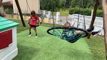 A child swinging on a net swing in a grassy outdoor area with a building in the background.