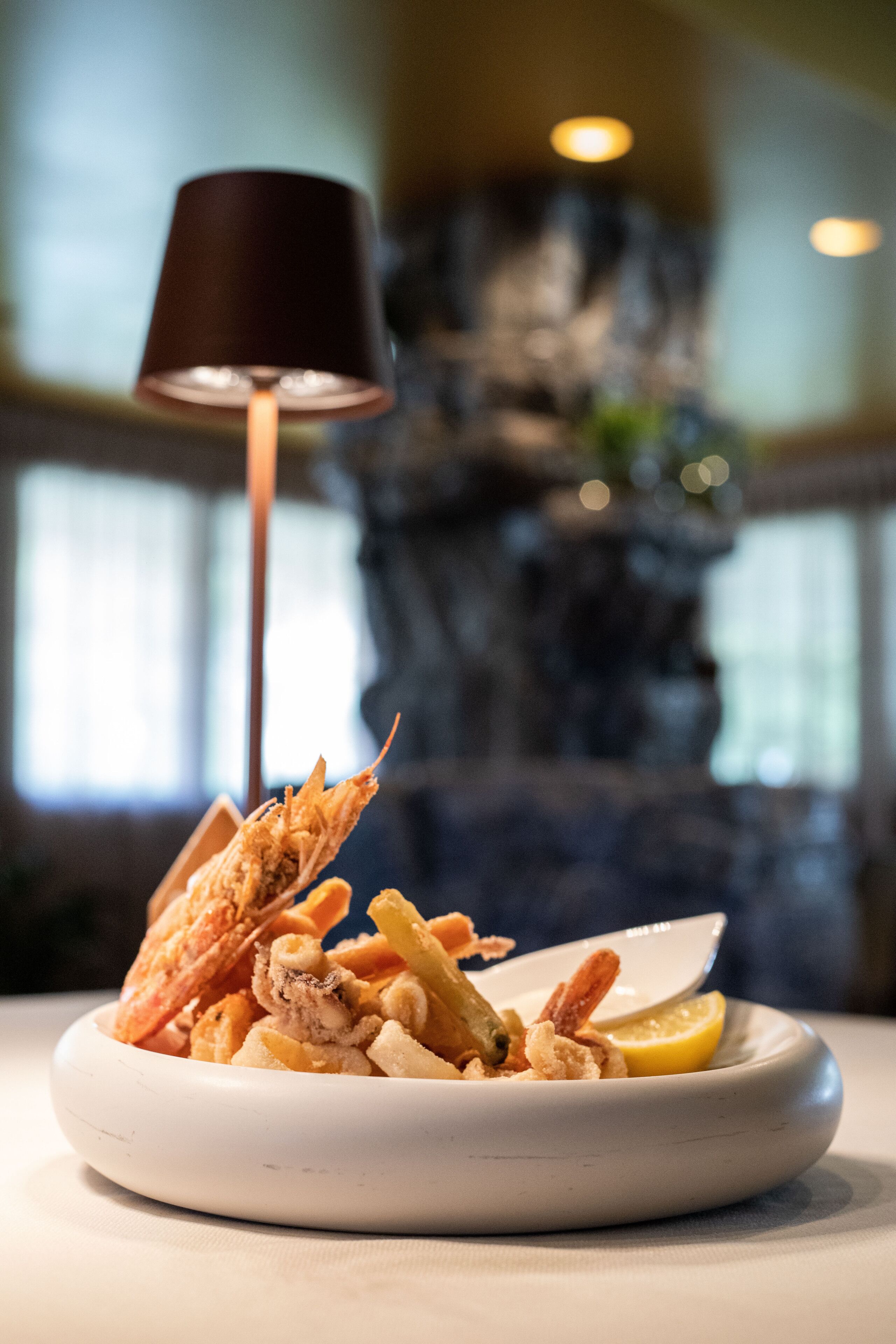 A plate of seafood with a lemon wedge and a small bowl of sauce, set on a table with a lamp in the background.