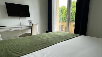 A hotel room with a bed, a desk, a chair, a flat-screen TV mounted on the wall, and a view of the outdoors through a large window.