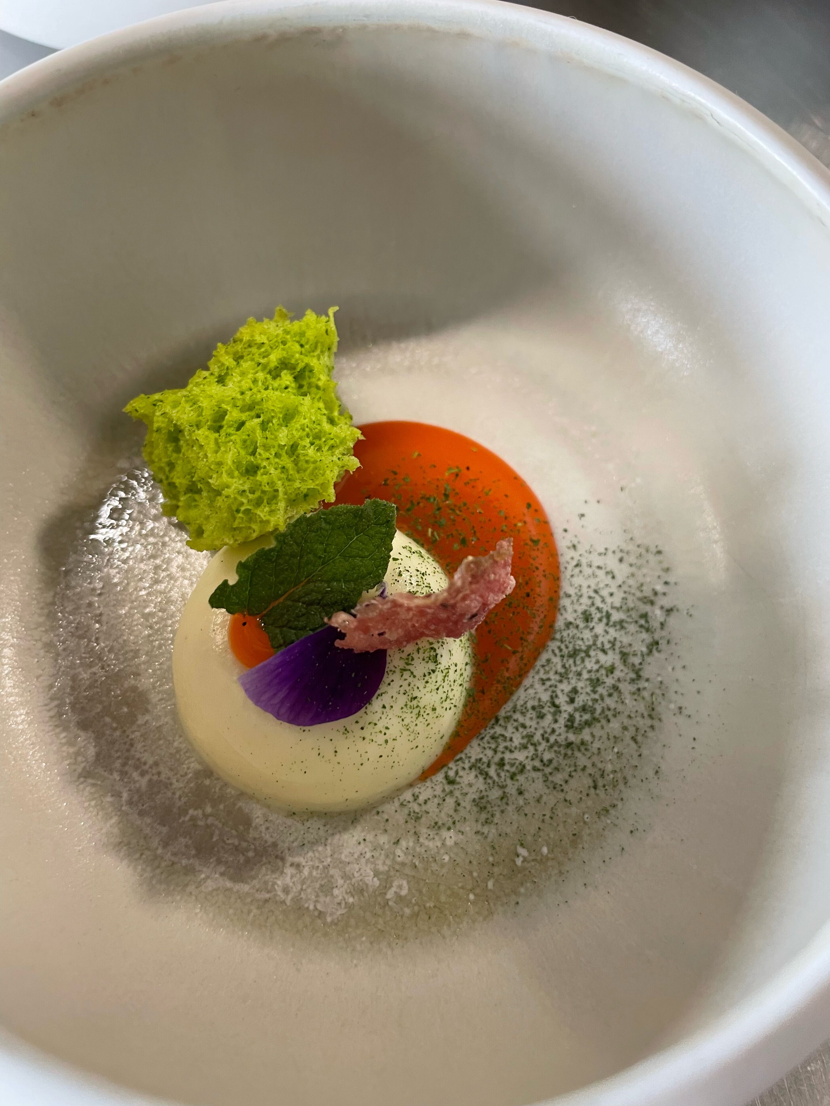 Un bol blanc amb un plat, una guarnició verda, una fulla morada i un element rosat, tot envoltat d'una salsa vermellosa.