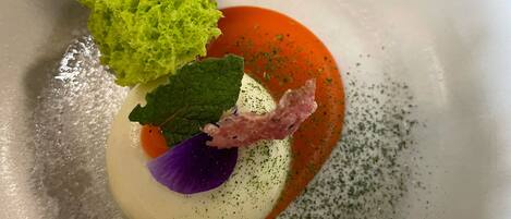 Un tazón blanco con un plato que tiene un adorno verde, una hoja morada y un elemento rosado, todo rodeado de salsa rojiza.