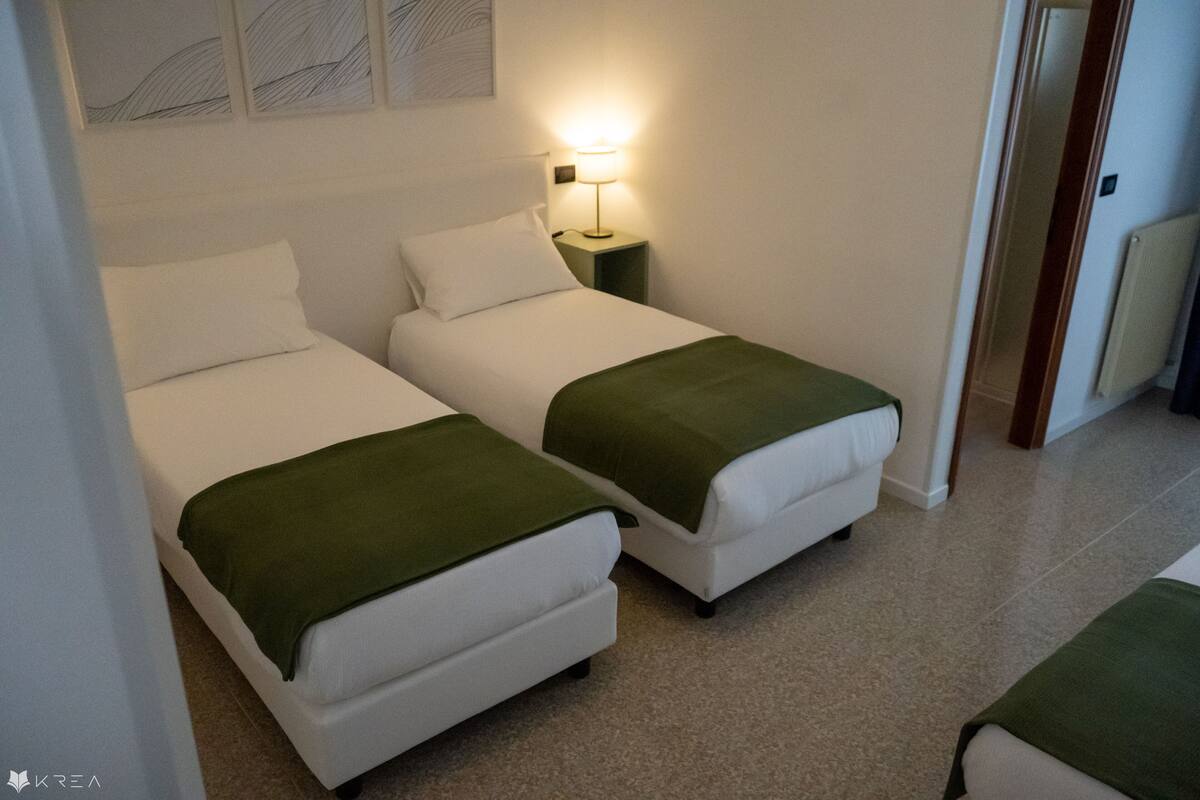 A room with two single beds, each with a green blanket, a bedside table with a lamp, and a wall-mounted artwork.