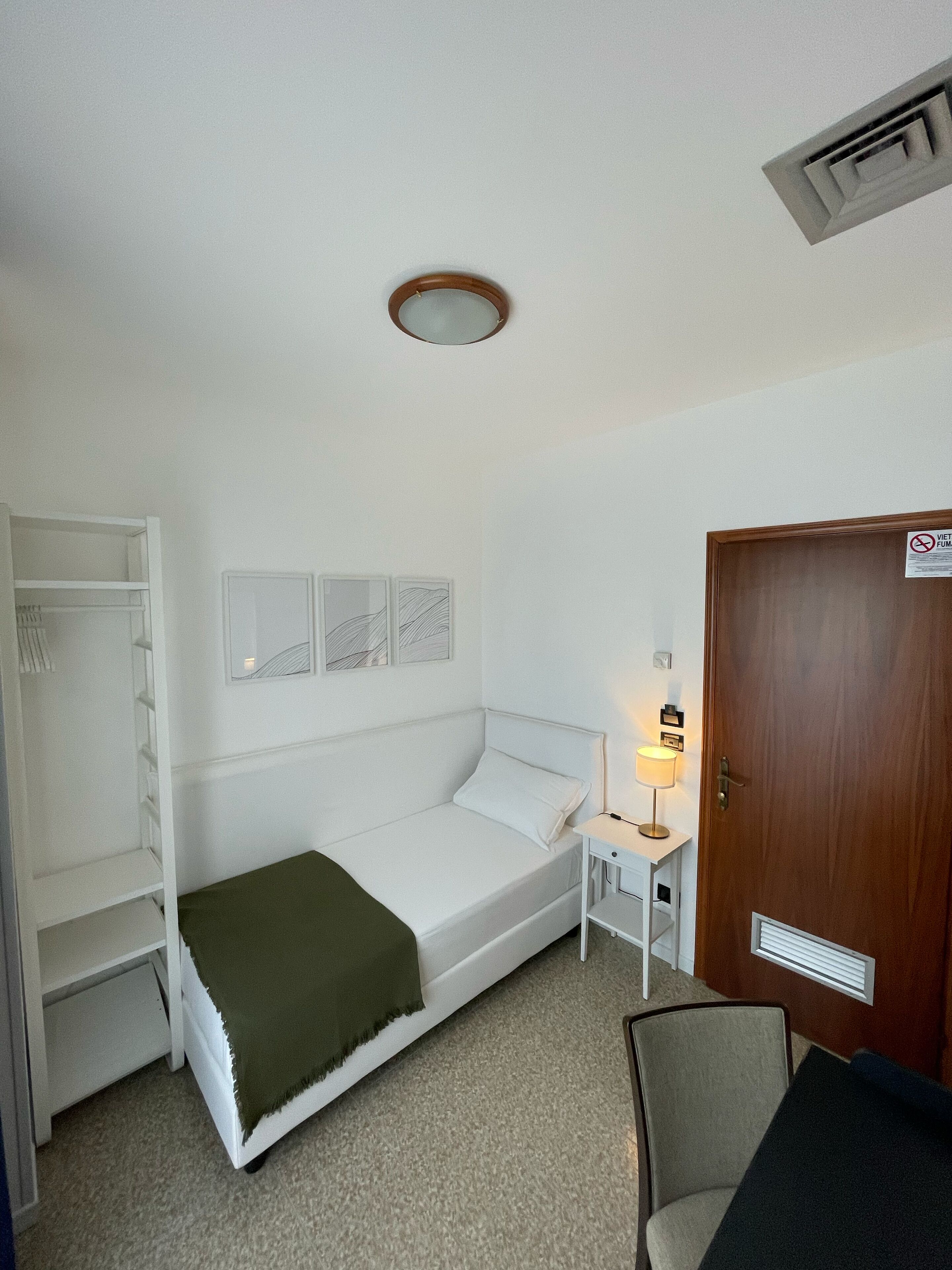 Una habitación pequeña con cama individual, una cama blanca, una mesita de noche con lámpara, una puerta de madera y un armario con estantes.