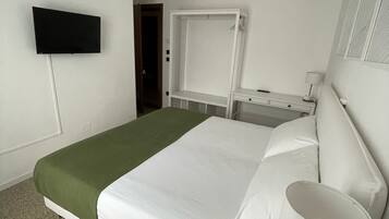 A single-bed room with a green bedspread, white bedding, a bedside table with a lamp, a TV mounted on the wall, and a window with blinds.