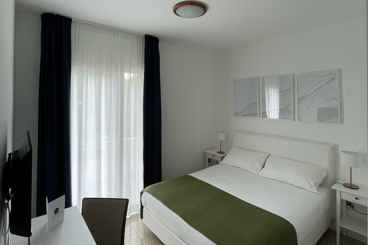 A hotel room with a bed, a desk, a chair, a nightstand, and a window with curtains.