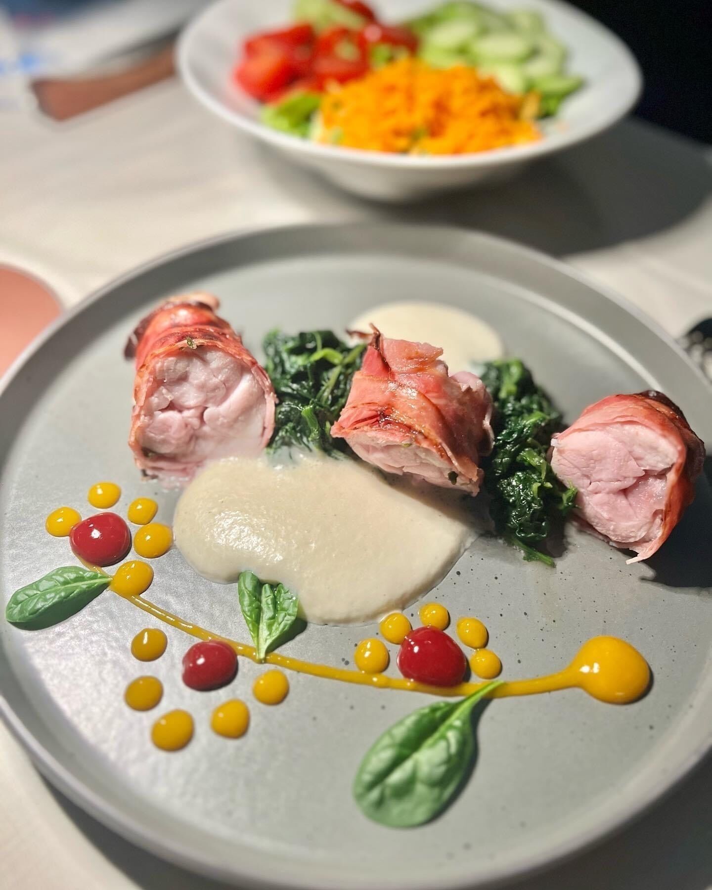 Une assiette de nourriture contenant du jambon, des légumes verts et de la sauce, et une autre assiette avec des légumes en arrière-plan.