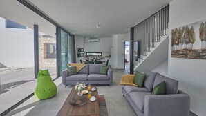 A modern living room with a grey sofa, green bean bag, and a coffee table with a vase of flowers. A staircase with a railing is visible in the background.