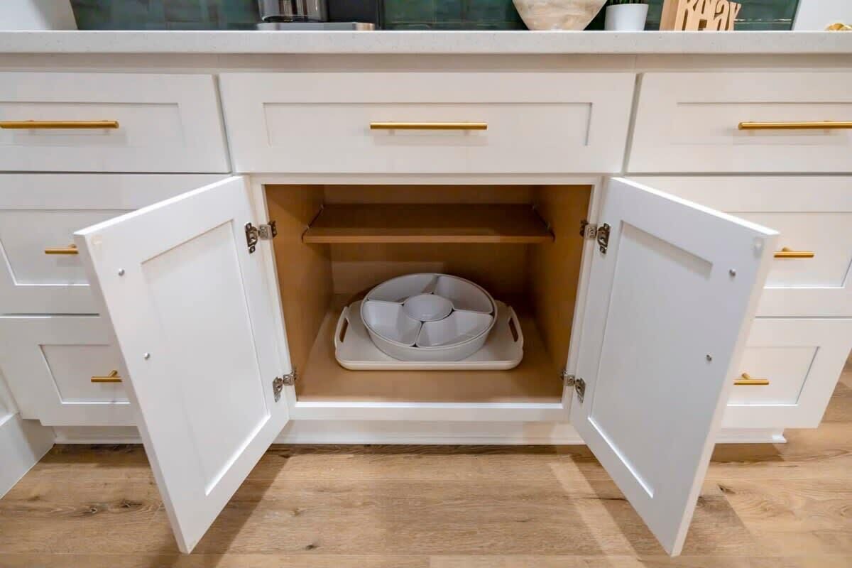 Ein weißes Schränkchen mit goldenen Griffen, eine geöffnete Tür zeigt ein weißes Tablett mit einem runden Gegenstand, und ein Holzboden.