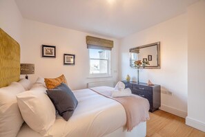 Un dormitorio con cama, mesita de noche, cómoda, ventana y espejo instalado en la pared.