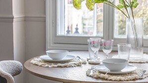 Una mesa redonda para comer con platos blancos, cristalería transparente y un jarrón con flores.
