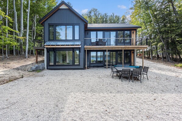 A modern two-story house with large windows, a deck, and a gravel driveway. - 🛶 Sunrise Sands Retreat | Torch Lakefront Home w/ Beach & Dock | Game Room ➠ 2364 (Kewadin)