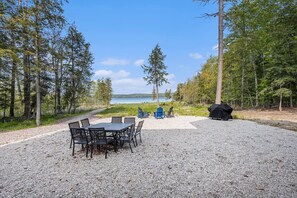 A gravel patio with a table and chairs, a covered grill, and Adirondack chairs. - 🛶 Sunrise Sands Retreat | Torch Lakefront Home w/ Beach & Dock | Game Room ➠ 2364 (Kewadin)